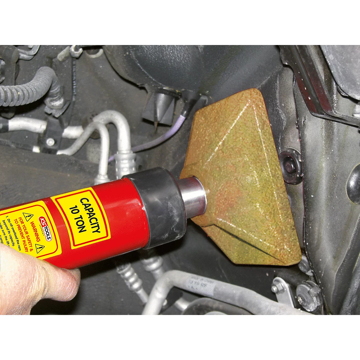 Ein Mann hält eine rote Hydraulikpresse mit der Aufschrift "CAPACITY 10 TON" an einem dreieckigen Hebeklotz im Motorraum eines Autos fest.